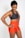 A smiling woman wearing a dark sports bra and bright orange athletic shorts with a logo on the side. She stands with her hands on her hips, exuding confidence and athleticism. Her hair is pulled back, and she seems to be in a positive mood.