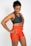 A smiling woman wearing a dark sports bra and bright orange athletic shorts with a logo on the side. She stands with her hands on her hips, exuding confidence and athleticism. Her hair is pulled back, and she seems to be in a positive mood.