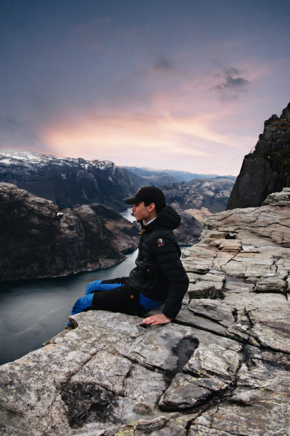 woman in black jacket and blue denim jeans sitting on rock formation near lake during daytime