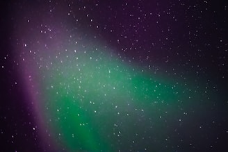 A celestial display of the northern lights with a vibrant mix of green and purple hues. The sky is dotted with numerous stars, creating a serene and awe-inspiring night scene.