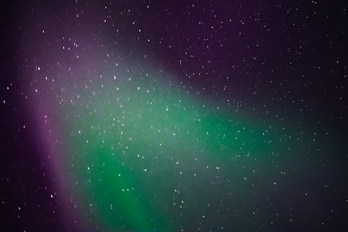 A celestial display of the northern lights with a vibrant mix of green and purple hues. The sky is dotted with numerous stars, creating a serene and awe-inspiring night scene.