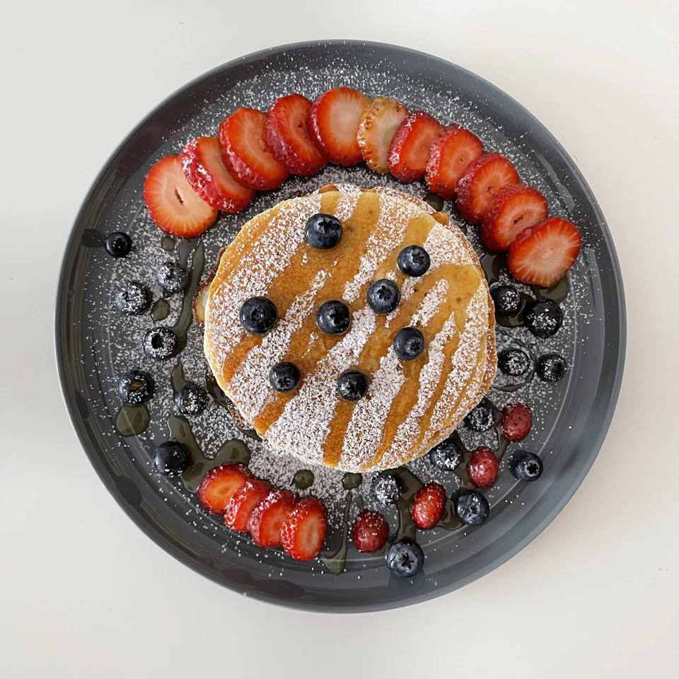 The image shows a round, dark gray plate with a white rim, containing a stack of pancakes topped with fresh blueberries and powdered sugar. The pancakes are generously drizzled with syrup, adding to their visual appeal. Scattered around the plate are additional blueberries and strawberries, providing a colorful contrast to the golden-brown pancakes.