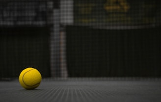 A close-up of a tennis ball resting on a vibrant neon yellow-green court surface, with a blurred net in the background.