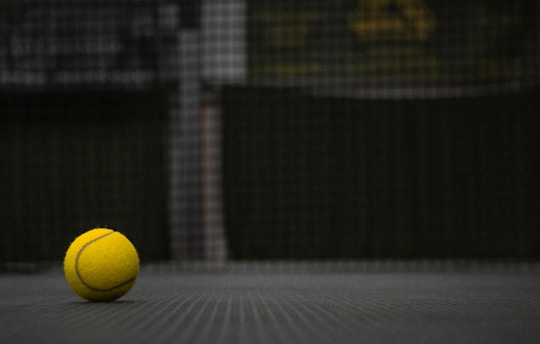 A close-up of a tennis ball resting on a vibrant neon yellow-green court surface, with a blurred net in the background.