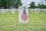 Rows of white gravestones are neatly arranged in a grassy cemetery. Small American flags are placed in front of each gravestone, indicating a tribute to veterans. Lush green trees are visible in the background under a partly cloudy sky.