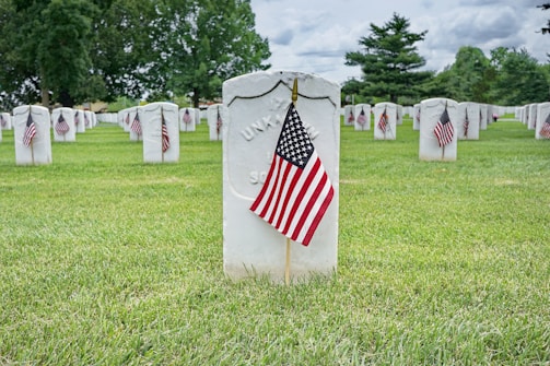 Rows of white gravestones are neatly arranged in a grassy cemetery. Small American flags are placed in front of each gravestone, indicating a tribute to veterans. Lush green trees are visible in the background under a partly cloudy sky.