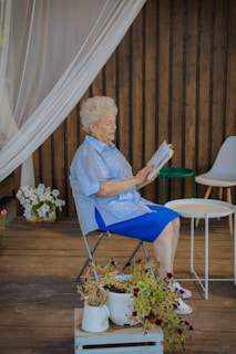 woman in blue dress shirt sitting on white folding chair reading book