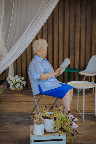 woman in blue dress shirt sitting on white folding chair reading book