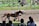 A horse race is taking place with a brown and a white horse racing neck and neck on a dirt track. A rider leans low as the horses gallop past spectators in the foreground. The track is bordered with colorful flags and banners. In the background, there is a green field with decorative structures and statues.