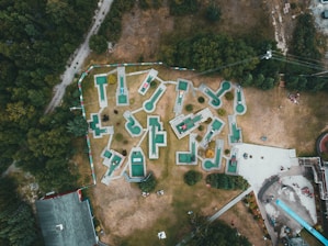 An aerial view of a mini-golf course with uniquely shaped holes spread across a grassy area surrounded by trees. The mini-golf holes are marked with green and white surfaces, connected by pathways. A few groups of people can be seen near the entrances and playing around the course. Adjacent to the golf course, there are buildings and a long slide, with additional people gathered around these areas.
