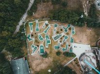An aerial view of a mini-golf course with uniquely shaped holes spread across a grassy area surrounded by trees. The mini-golf holes are marked with green and white surfaces, connected by pathways. A few groups of people can be seen near the entrances and playing around the course. Adjacent to the golf course, there are buildings and a long slide, with additional people gathered around these areas.