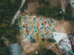 An aerial view of a mini-golf course with uniquely shaped holes spread across a grassy area surrounded by trees. The mini-golf holes are marked with green and white surfaces, connected by pathways. A few groups of people can be seen near the entrances and playing around the course. Adjacent to the golf course, there are buildings and a long slide, with additional people gathered around these areas.