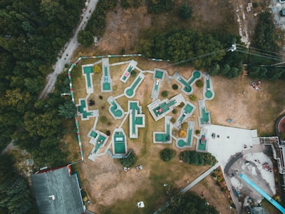 An aerial view of a mini-golf course with uniquely shaped holes spread across a grassy area surrounded by trees. The mini-golf holes are marked with green and white surfaces, connected by pathways. A few groups of people can be seen near the entrances and playing around the course. Adjacent to the golf course, there are buildings and a long slide, with additional people gathered around these areas.