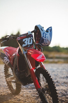 A red Honda dirt bike parked on a rugged, pebbled terrain. The bike is adorned with a number 30 racing decal and a blue helmet is casually placed on the handlebars. The background is a blurred view of natural greenery under a clear sky, suggesting an outdoor setting.