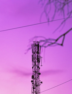 A telecommunications tower is silhouetted against a vibrant purple sky. Some faint shadows of tree branches are visible in the foreground, giving a dynamic frame to the composition. The image features electrical wires stretching across the sky.