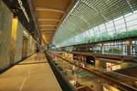 A spacious, modern shopping mall interior with a high, arched glass ceiling allowing natural light to flood in. Multiple levels of retail stores are visible, with sleek, reflective floors and contemporary design elements. The upper level features greenery along the side, creating a vibrant, inviting atmosphere. Shoppers are casually walking along the corridors, contributing to a lively but relaxed environment.
