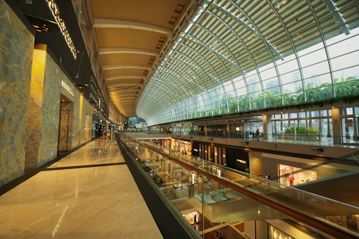 A spacious, modern shopping mall interior with a high, arched glass ceiling allowing natural light to flood in. Multiple levels of retail stores are visible, with sleek, reflective floors and contemporary design elements. The upper level features greenery along the side, creating a vibrant, inviting atmosphere. Shoppers are casually walking along the corridors, contributing to a lively but relaxed environment.