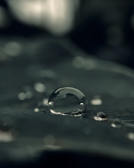 A close-up view of a large water droplet resting on a dark, smooth surface. Smaller droplets are scattered around the larger one, and the background is blurred, emphasizing the droplet's clarity and reflective qualities.