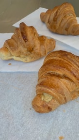 Three croissants are placed on white parchment paper, showcasing a flaky, golden-brown crust. One of the croissants has a filling visible at the end. The croissants appear freshly baked and appetizing.