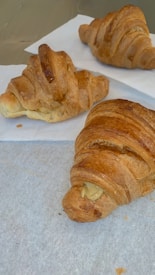 Three croissants are placed on white parchment paper, showcasing a flaky, golden-brown crust. One of the croissants has a filling visible at the end. The croissants appear freshly baked and appetizing.