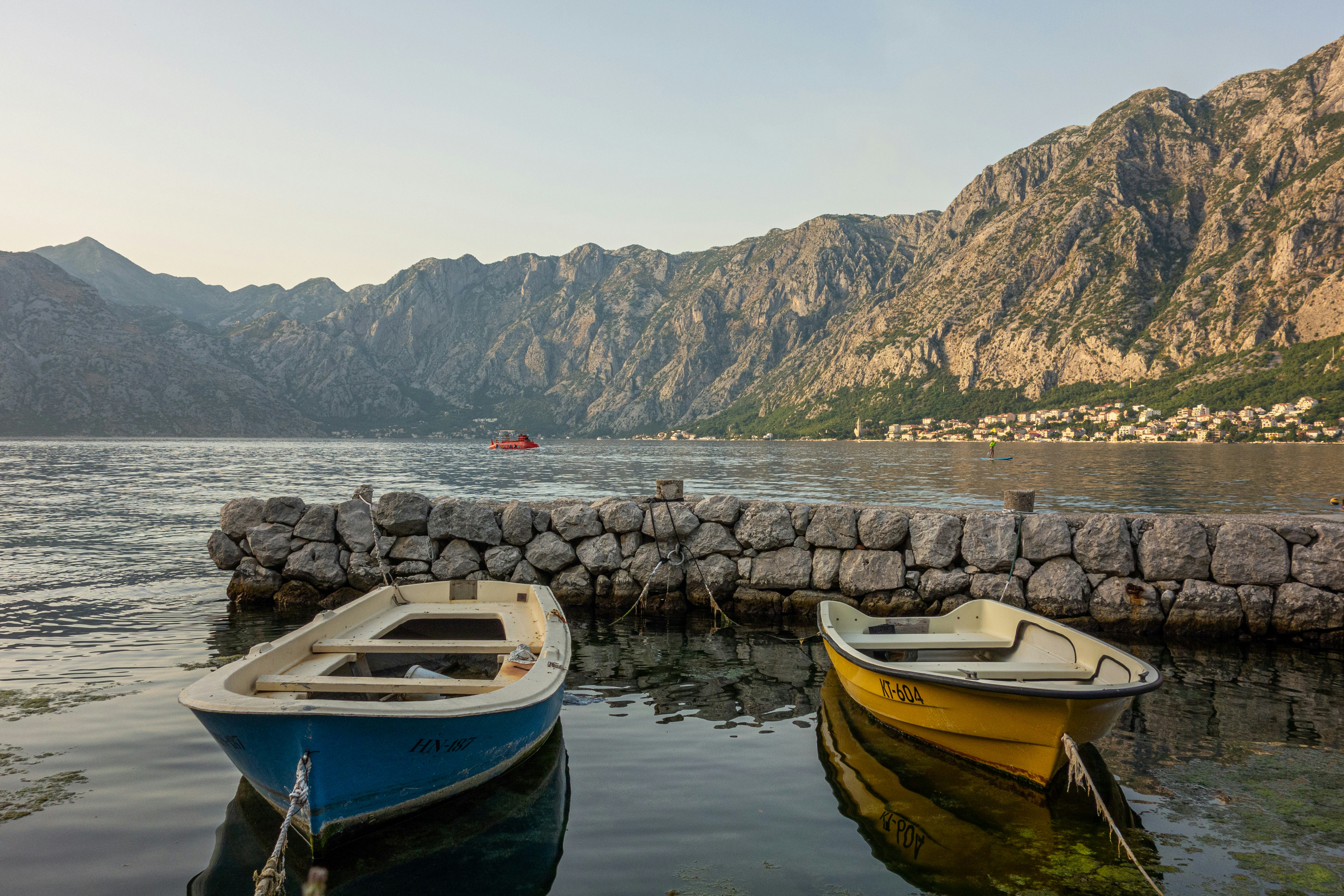 Two small fishing boats moored at a stone breakwater on the clear, calm Bay of Kotor with the mountains rising behind