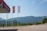 A view of a gas station with the sign 'Kalamper Petro' prominently displayed. Three flags are flying, one with a red background and emblem, and two with the gas station's branding. In the background, a range of green-covered mountains stretches across under a clear blue sky. The foreground features a paved area and some greenery.