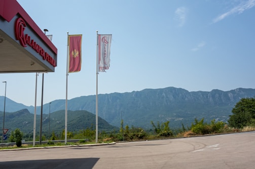 A view of a gas station with the sign 'Kalamper Petro' prominently displayed. Three flags are flying, one with a red background and emblem, and two with the gas station's branding. In the background, a range of green-covered mountains stretches across under a clear blue sky. The foreground features a paved area and some greenery.