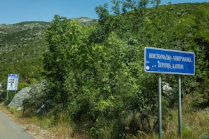 A country road is bordered by lush greenery and shrubs. Two blue road signs are present; one is close to the foreground with the text 'Hercegovačko-Neretvanska Županija-Kanton', and another smaller sign is further down the road, displaying various symbols. The background consists of mountainous terrain under a clear blue sky.