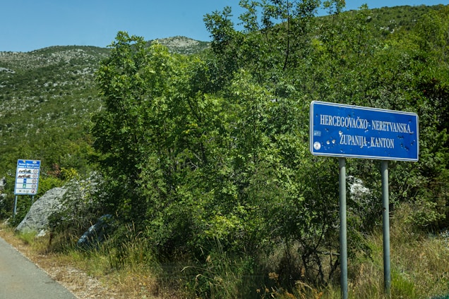 A country road is bordered by lush greenery and shrubs. Two blue road signs are present; one is close to the foreground with the text 'Hercegovačko-Neretvanska Županija-Kanton', and another smaller sign is further down the road, displaying various symbols. The background consists of mountainous terrain under a clear blue sky.
