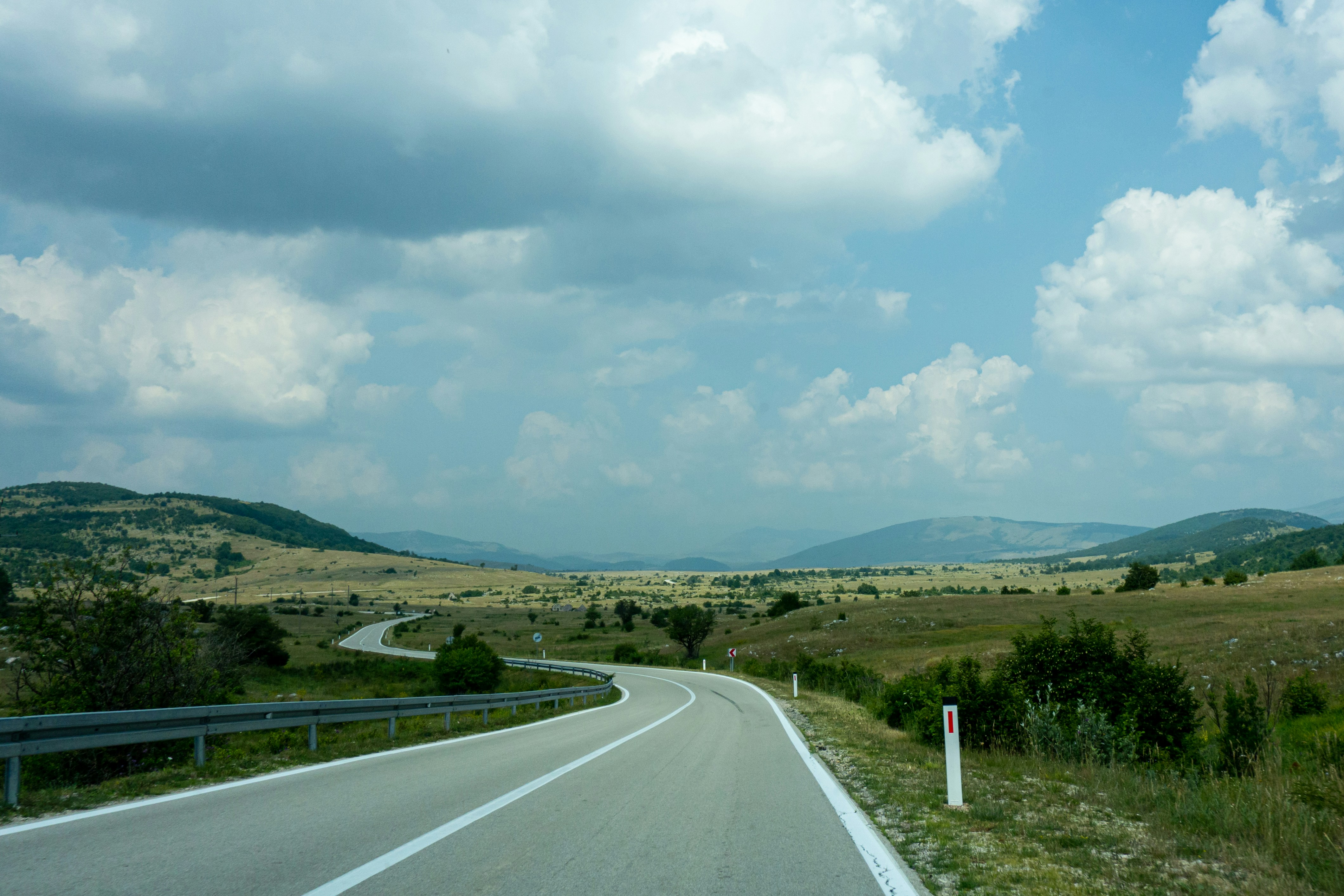 gray concrete road between green grass field under white clouds and blue sky during daytime