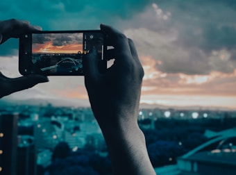 A person holds a smartphone horizontally, capturing a photograph of a vibrant sunset over a cityscape. The phone screen displays a clear image of a city with a sky painted in warm tones of orange and yellow, contrasting with the darker shadows of the buildings. The background includes a panoramic view of the city under a blue sky with thick clouds.