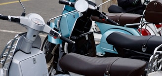 A close-up view of multiple parked scooters, showcasing a variety of colors and designs. The scooters have sleek and modern designs, featuring mirrors, seats, and handlebars. The setting appears to be an outdoor parking area.