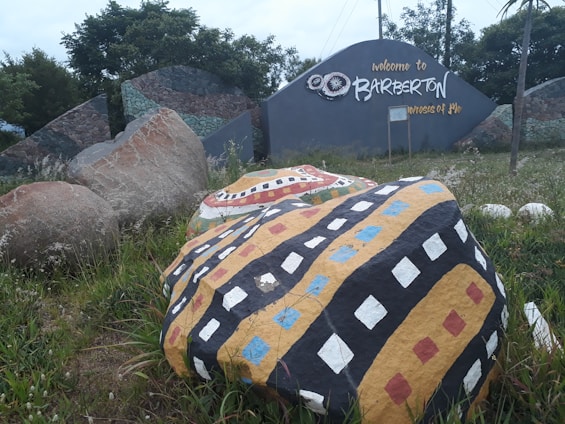 Colorfully painted rocks with geometric patterns are surrounded by greenery. The scene also features a large sign welcoming visitors to Barberton, hinting at a cultural or tourist landmark. Broad stones with pebble mosaics form part of the backdrop.