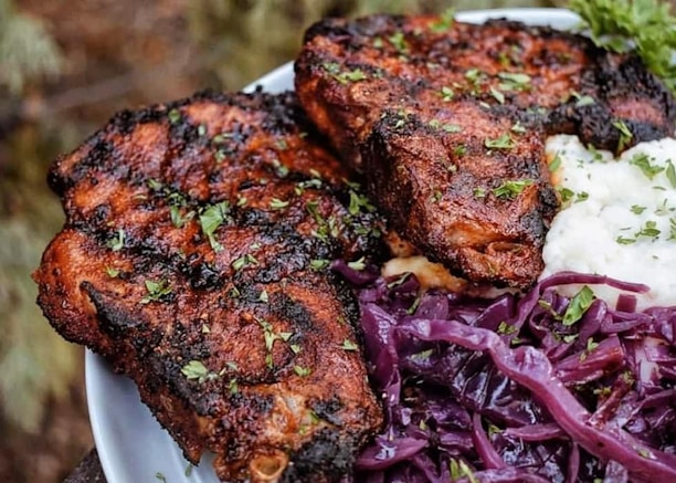 Grilled meat seasoned with herbs, placed on a plate beside vibrant purple cabbage and a portion of creamy mashed potatoes garnished with fresh green parsley.
