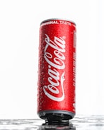 A vertical red can of Coca-Cola with water droplets on its surface. The words 'Coca-Cola' and 'Original Taste' are prominently displayed. The can is set against a white background with a reflective surface underneath it.