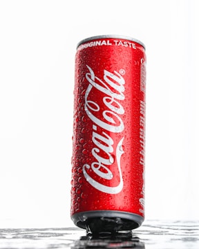 A vertical red can of Coca-Cola with water droplets on its surface. The words 'Coca-Cola' and 'Original Taste' are prominently displayed. The can is set against a white background with a reflective surface underneath it.