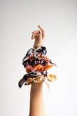 A raised hand with a light skin tone adorned with multiple scrunchies in various colors and patterns is depicted against a plain, light-colored background. The scrunchies feature vibrant and contrasting prints, including floral designs and abstract patterns.