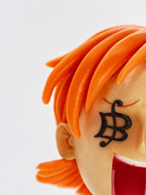 A close-up of a figurine with vibrant orange hair and a unique black symbol over its eye. The figure appears to be stylized with exaggerated features, including a wide-open mouth.