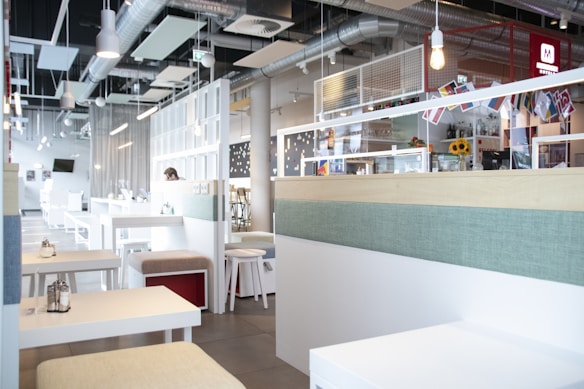 A modern, bright cafe interior with sleek white furniture and tables, complemented by pastel-colored cushions. The ceiling features exposed ducts and industrial-style lighting. Decorative items and pictures adorn the walls, and a counter with colorful flags and sunflowers is visible. The space appears spacious and tidy, with a calm atmosphere.