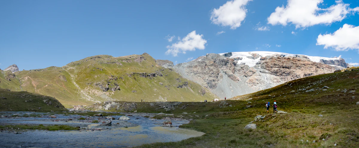 Hikers by a stream in the Vallone delle Cime Bianche, under a snow-capped peak