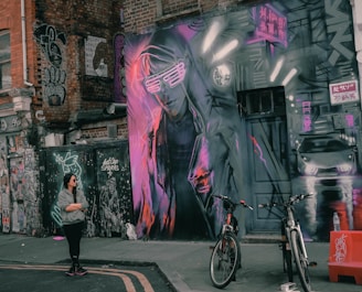 A vibrant street art scene with graffiti covering a brick building and a large mural depicting a futuristic figure wearing neon sunglasses. The wall has various neon-colored elements, including signs written in an East Asian script and images of a car. A woman stands on the street observing the art, and two bicycles are parked nearby.
