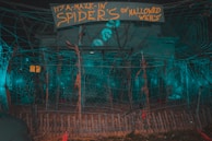 A scene depicting a spooky outdoor display with large spider webs spread across a fenced area. A sign hangs above with playful text, adding to the Halloween theme. The area is dimly lit with a blue-green hue, creating an eerie atmosphere.