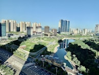 A modern urban landscape featuring a densely built area with high-rise buildings surrounded by a meticulously landscaped park and a central waterway. The foreground shows wide roads with minimal traffic, and the scene is illuminated by bright daylight.