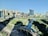 A modern urban landscape featuring a densely built area with high-rise buildings surrounded by a meticulously landscaped park and a central waterway. The foreground shows wide roads with minimal traffic, and the scene is illuminated by bright daylight.