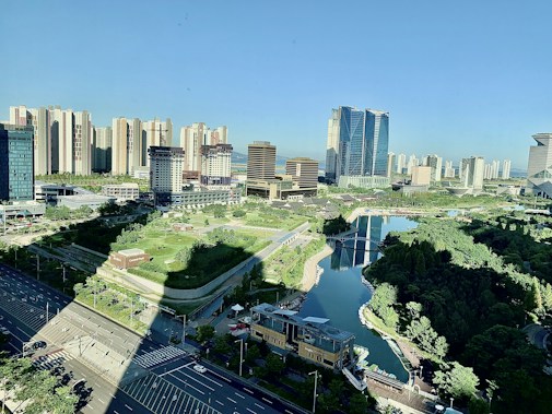 A modern urban landscape featuring a densely built area with high-rise buildings surrounded by a meticulously landscaped park and a central waterway. The foreground shows wide roads with minimal traffic, and the scene is illuminated by bright daylight.