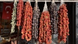 A display of various dried vegetables hangs in bunches outside a shop. The vegetables include peppers and eggplants. The scene is set against the backdrop of a Turkish flag visible inside the store, indicating cultural and geographic context.