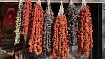 A display of various dried vegetables hangs in bunches outside a shop. The vegetables include peppers and eggplants. The scene is set against the backdrop of a Turkish flag visible inside the store, indicating cultural and geographic context.