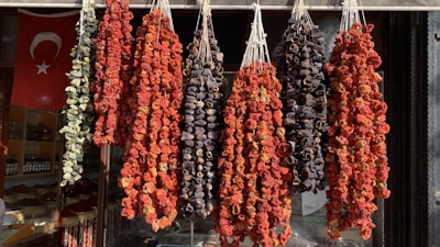 A display of various dried vegetables hangs in bunches outside a shop. The vegetables include peppers and eggplants. The scene is set against the backdrop of a Turkish flag visible inside the store, indicating cultural and geographic context.