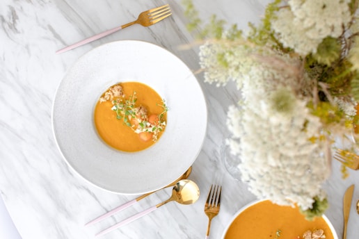 A beautifully presented bowl of orange soup garnished with microgreens and small round ingredients, resting on a white marble table. A pink-handled fork and gold spoon are placed nearby. There is a bouquet of white flowers slightly out of focus at the top right.