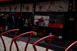 A group of people stand outside a store with signage in different languages and pictures of meat products. The store seems to specialize in various meats as indicated by the signs with illustrations. The entrance of the store is visible with some people interacting near it, possibly shopping or waiting.